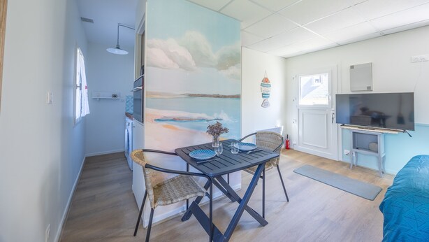 A compact living space with a dining area featuring a table and chairs, a kitchenette, a TV, and a large window. - Marée Basse - Studio proche plage de la potinière (Barneville-Carteret)