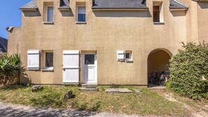 A two-story house with a beige exterior, a front door, two windows with shutters, a garage door, and a bicycle parked outside. - Marée Basse - Studio proche plage de la potinière (Barneville-Carteret)