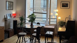 Un coin repas avec une table ronde en verre, quatre chaises, une plante en pot et une vue sur un bâtiment en briques, visible à travers de grandes fenêtres.