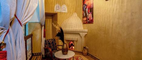 A cozy room with a patterned rug, a small round table, a decorative vase, and a wall-mounted shelf with a painting.