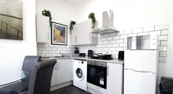 A compact kitchen with a washing machine, oven, microwave, and a dining area with a glass table and chair.