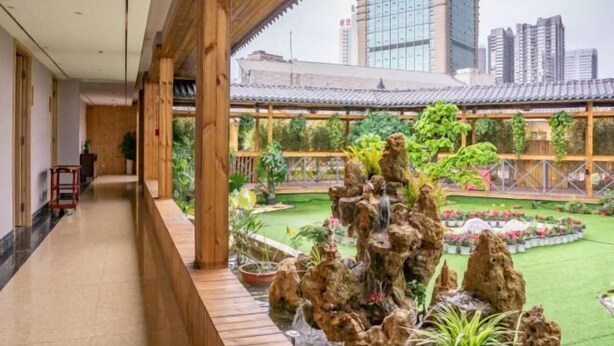 A hotel corridor with a wooden ceiling, a small indoor garden with a rock waterfall, and a view of the cityscape in the background.