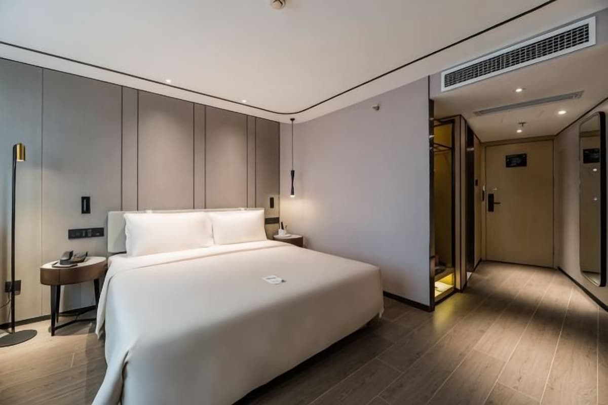 A modern hotel room with a large bed, bedside tables, a nightstand, a wall-mounted lamp, and a door leading to another room.