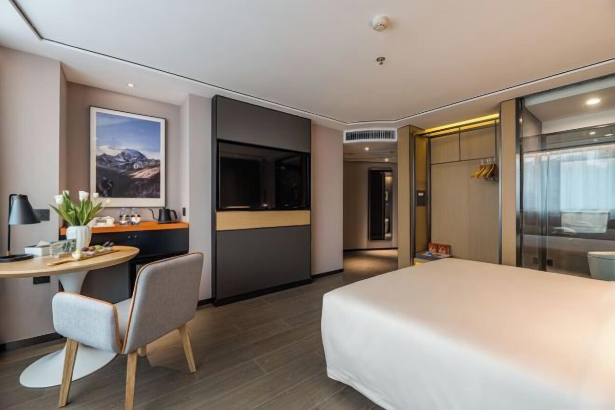Ein modernes Hotelzimmer mit Bett, Schreibtisch mit Stuhl, einem kleinen Tisch mit Pflanze und einem großen Fenster.