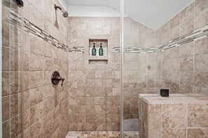A modern bathroom with beige tiles, a glass shower enclosure, and a small shelf with toiletries. - Historic Downtown Home with Fire Pit (Leesburg)