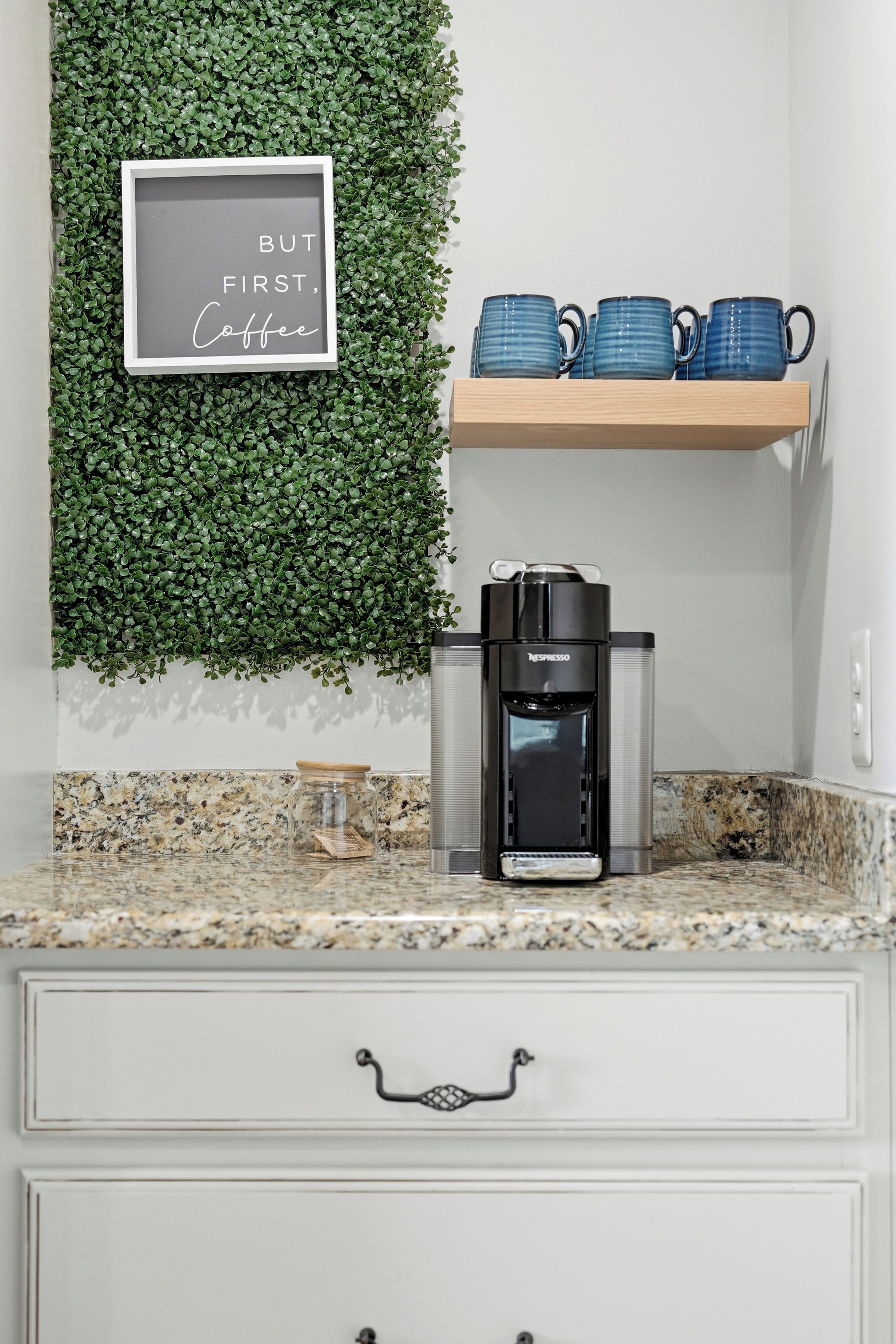 Une cuisine équipée d’une machine à café, d’un panneau décoratif encadré sur lequel est écrit : « D’abord, un café », et d’une étagère remplie de tasses bleues.