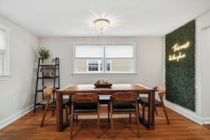 Un comedor con mesa y sillas de madera, una araña, un letrero de neón y una pared decorada con follaje verde.