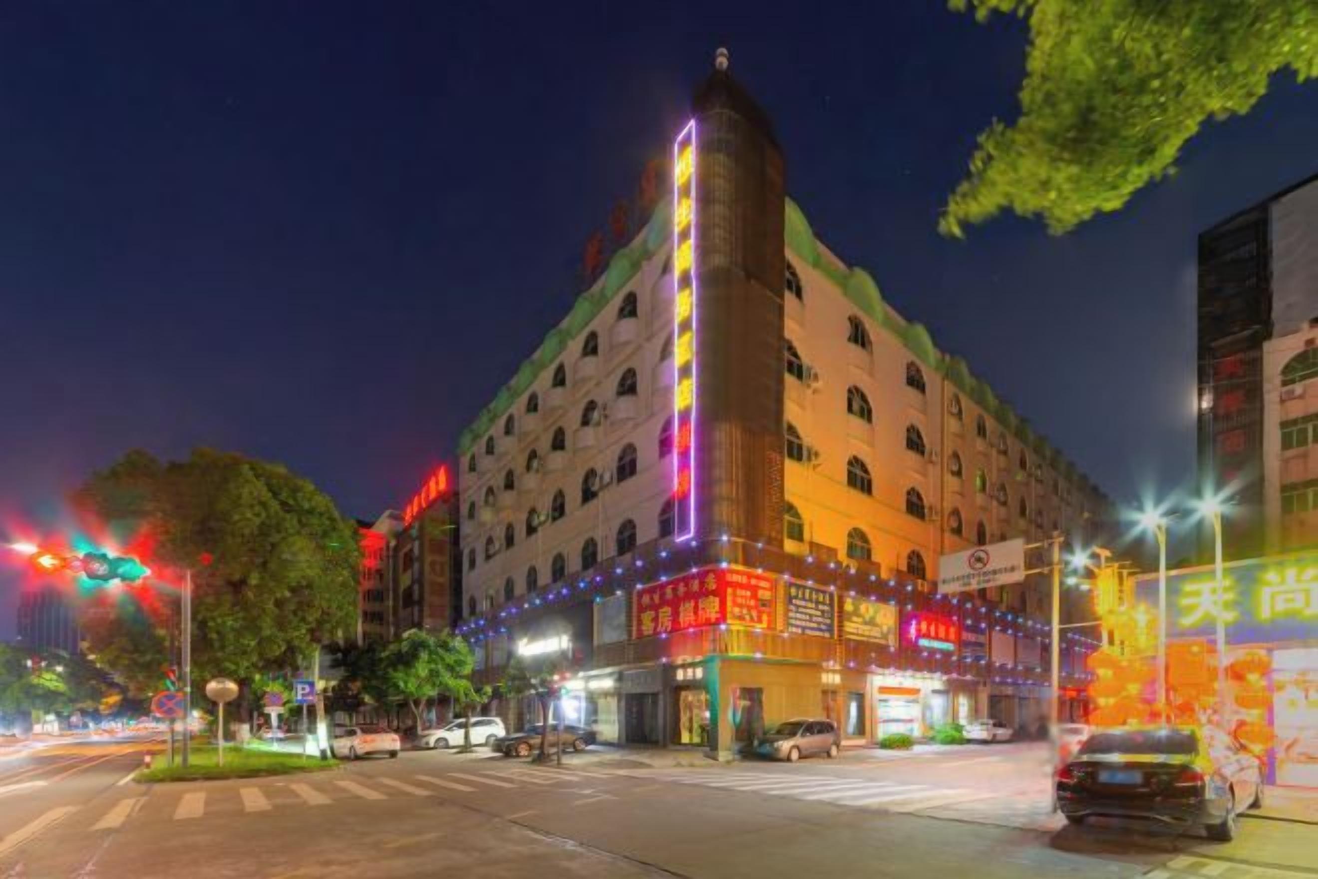 A multi-story hotel with a prominent corner tower, illuminated signage, and a street view with vehicles and trees.