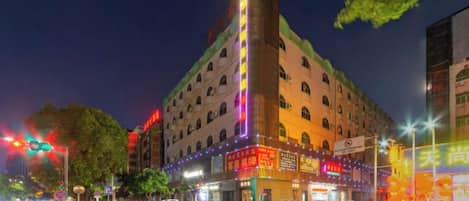 A multi-story hotel with a prominent corner tower, illuminated signage, and a street view with vehicles and trees.