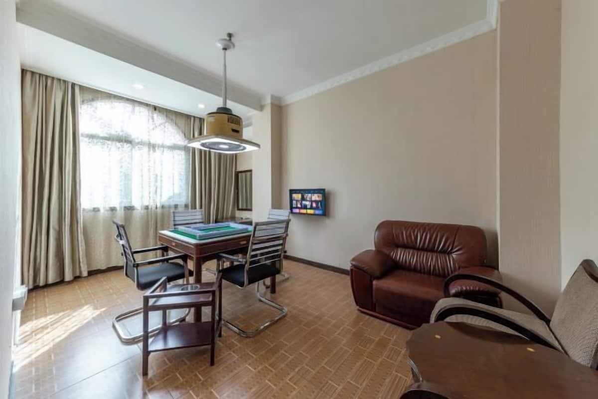 Een hotelkamer met een eettafel, stoelen, een bruine leren bank, een aan de muur bevestigde televisie en een groot raam met gordijnen.