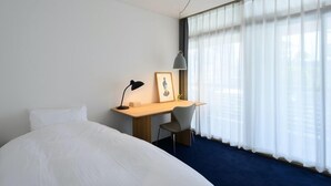 Une chambre d’hôtel avec un grand lit, un bureau avec une chaise, une lampe et une fenêtre avec des rideaux légers.