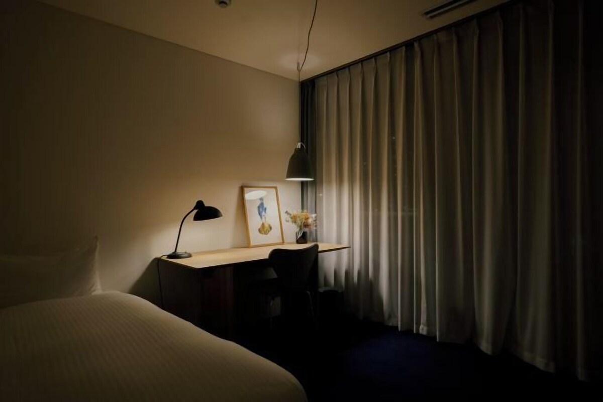 Une chambre d’hôtel avec un lit, un bureau, une chaise, une lampe et une fenêtre avec des rideaux.