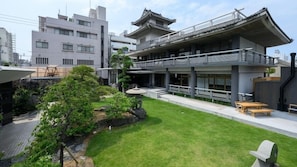 Un edificio moderno con tejado de estilo japonés tradicional, un jardín cuidado con césped y rocas, y una zona de estar con mesas y bancos.