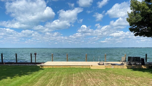 Eine Holzterrasse mit Rasenfläche, einem Metallgrill und Blick auf das Wasser.