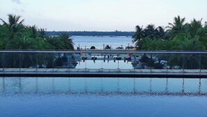 Vista nítida de uma piscina com barreira de vidro, cercada por palmeiras e uma vista distante do mar.