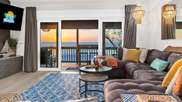 Un salon comprenant un grand canapé, une table basse et un téléviseur fixé au mur. Il y a un balcon d’où l’on peut admirer le coucher de soleil et l’océan.