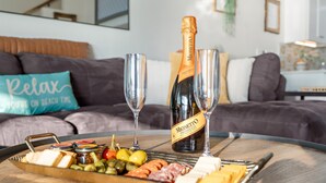 Une planche de charcuterie garnie de différents fromages, de viandes et d’olives, une bouteille de Prosecco Monetto et deux verres de champagne, le tout posé sur une table en bois dans un salon doté d’un canapé gris et d’un coussin orné du mot « Détente » à l’arrière-plan.