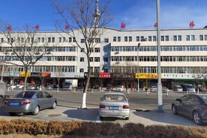 A multi-story building with commercial establishments on the ground floor, a parking area with cars, and a clear sky. - Jinshan Hotel (Harqin Banner)