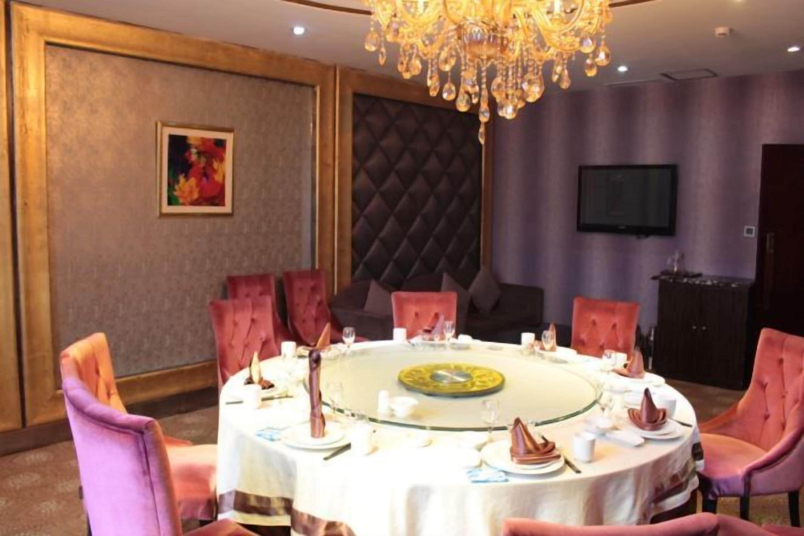 A round dining table with pink upholstered chairs, a chandelier, and a television on the wall.