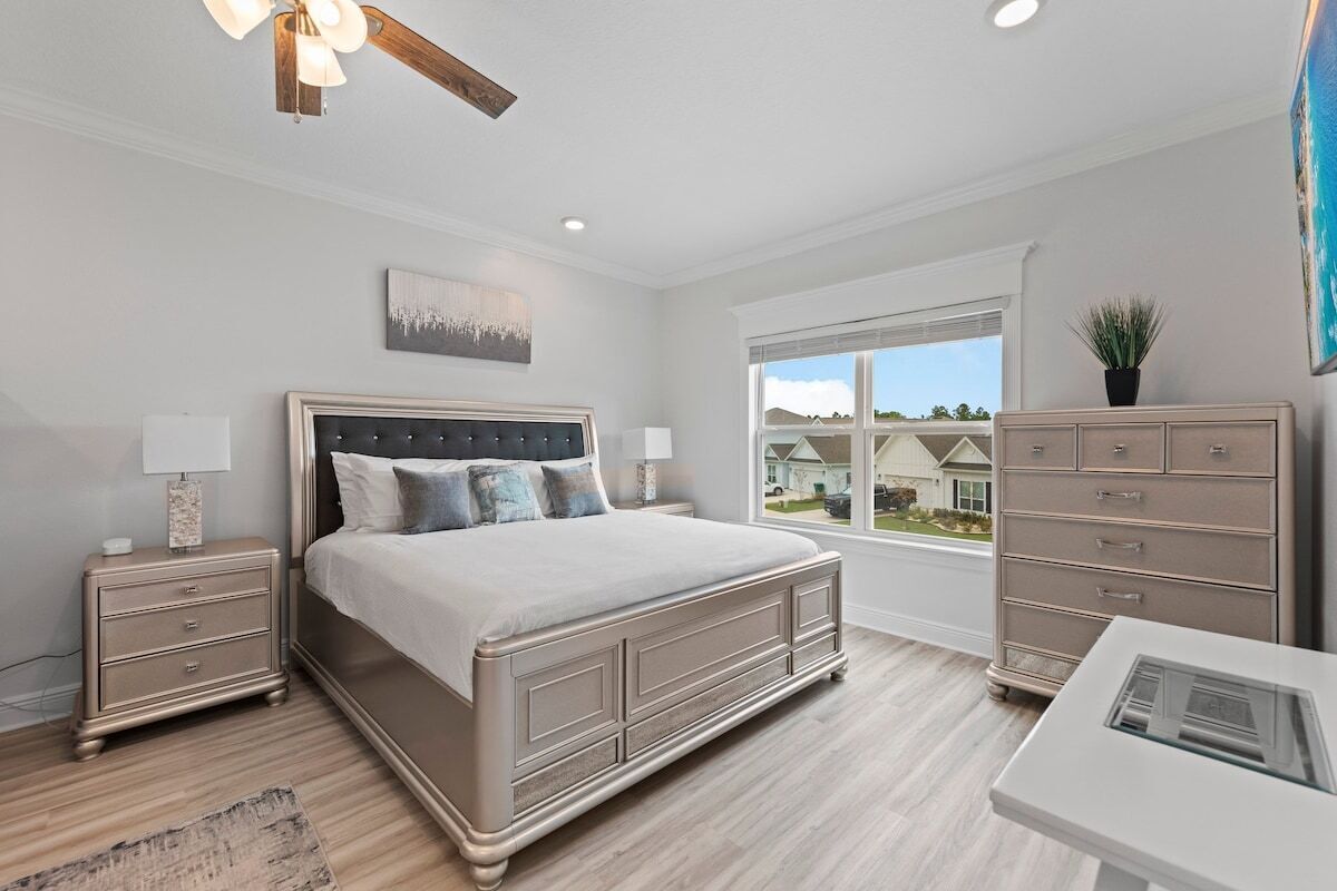 A bedroom with a large bed, bedside tables, a dresser, a ceiling fan, and a window with a view of houses.