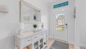 A white entryway with a door labeled "COASTAL HEAVEN," a mirror, a small table with a lamp, and a patterned rug. - 14 Beds | Sleeps 16 | Piper Cove Retreat | Pool | Santa Rosa Beach (Santa Rosa Beach)