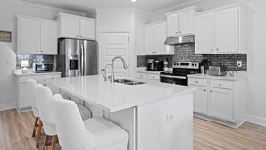 A modern kitchen with white cabinetry, a stainless steel refrigerator, a built-in oven, and a large island with seating. - 14 Beds | Sleeps 16 | Piper Cove Retreat | Pool | Santa Rosa Beach (Santa Rosa Beach)