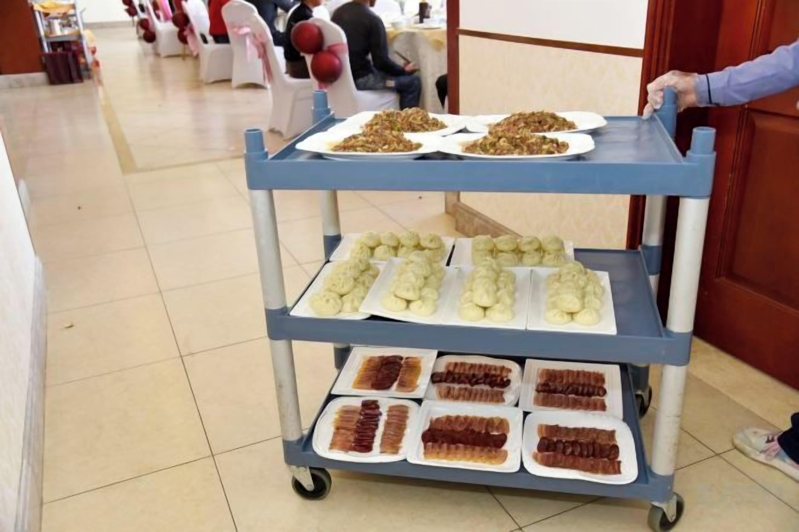A food cart with trays of various dishes, including what appears to be pastries and desserts, in a room with a tiled floor and a door in the background.