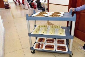 Carrinho de comida com bandejas de diversos pratos, incluindo aparentemente doces e sobremesas, em um espaço com piso ladrilhado e uma porta ao fundo.