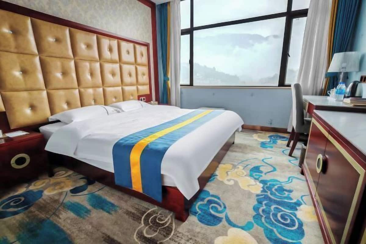 Habitación de hotel con una cama grande, cabecera de madera, escritorio con silla, ventana con vistas y alfombra estampada.