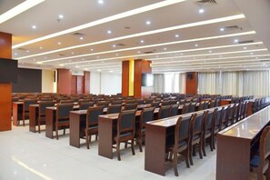 Uma sala de conferências com fileiras de cadeiras e mesas, uma TV de tela plana e um grande janelão com cortinas.
