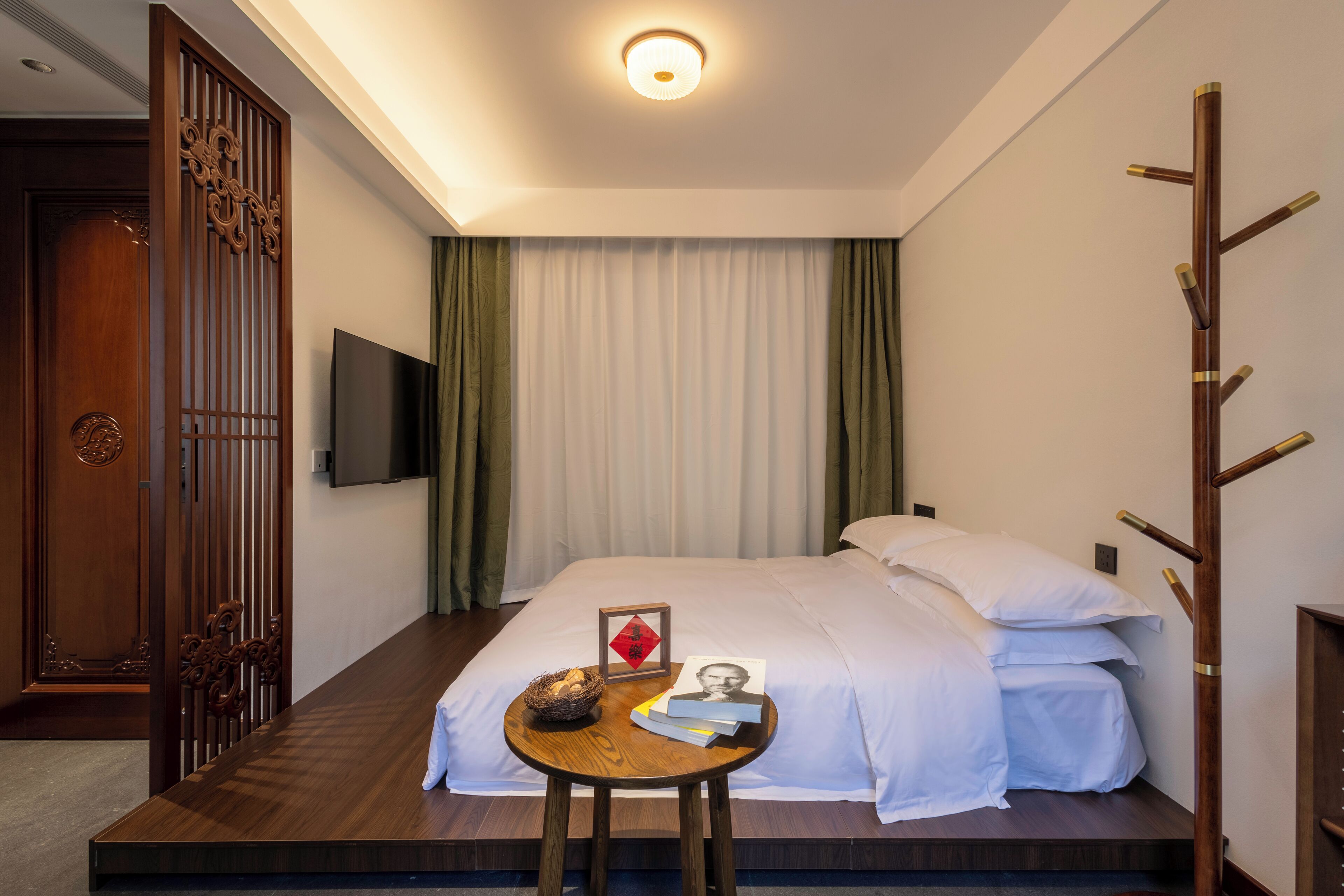 A hotel room with a bed, a small table with books and a picture frame, a TV on the wall, and a wooden partition.