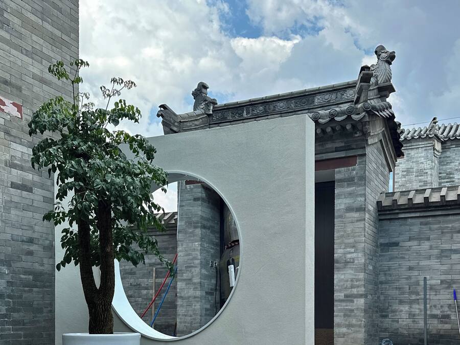 Pátio com árvore em vaso, espelho circular e edifício tradicional com telhado de azulejos.