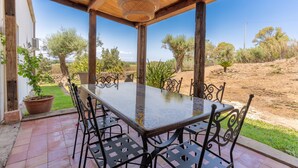 A covered outdoor dining area with a table and chairs, surrounded by potted plants and a view of the countryside.