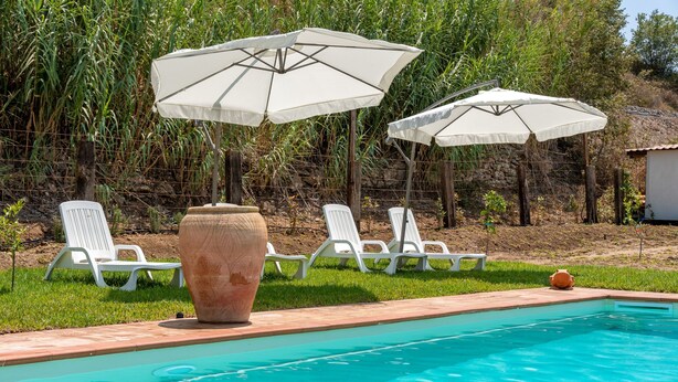 A swimming pool with white lounge chairs and umbrellas, surrounded by greenery and a stone wall.