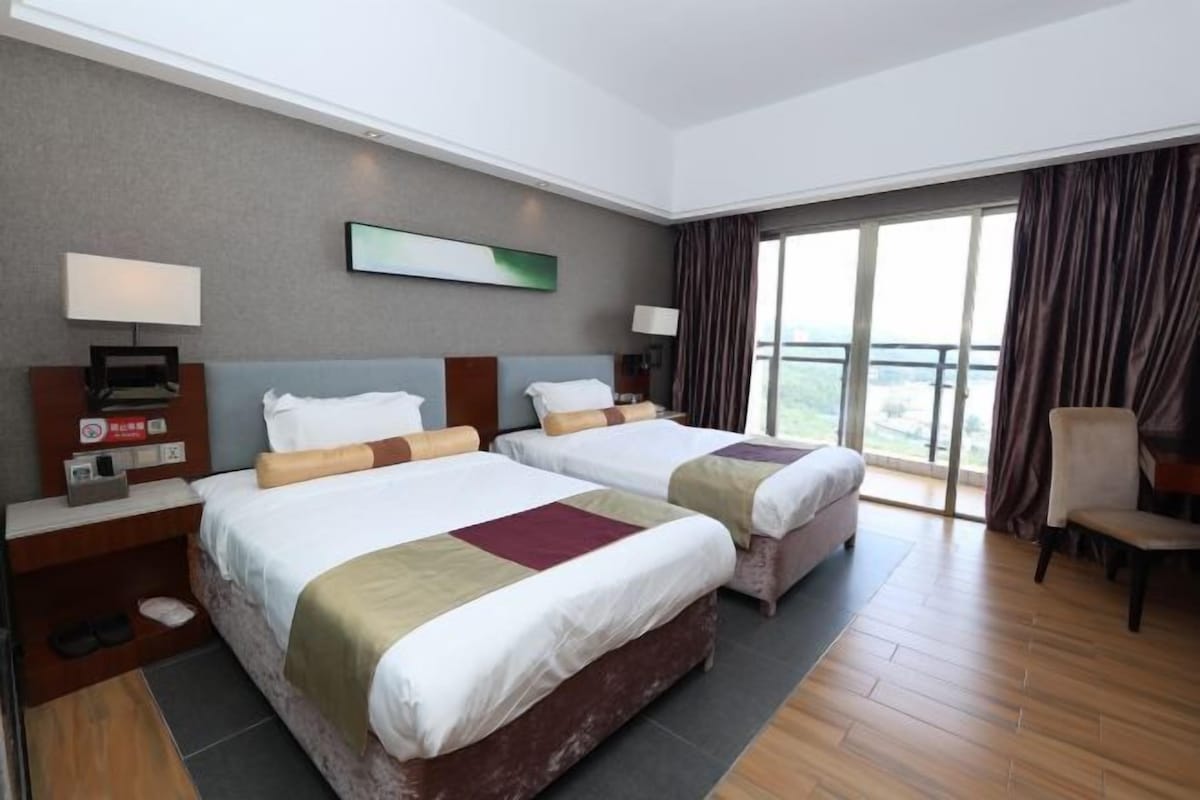 A hotel room with two beds, a large window with curtains, a chair, and a view of the outdoors.