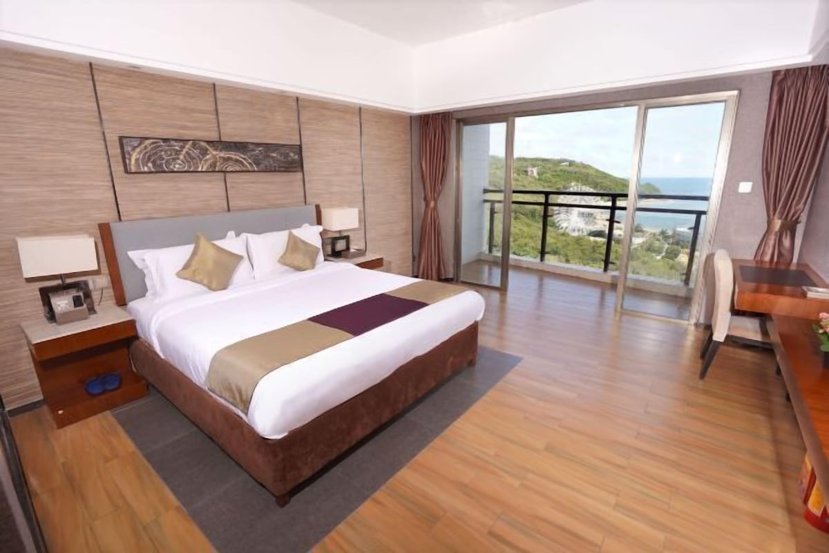 A hotel room with a large bed, a nightstand, a desk, and a balcony with a view.
