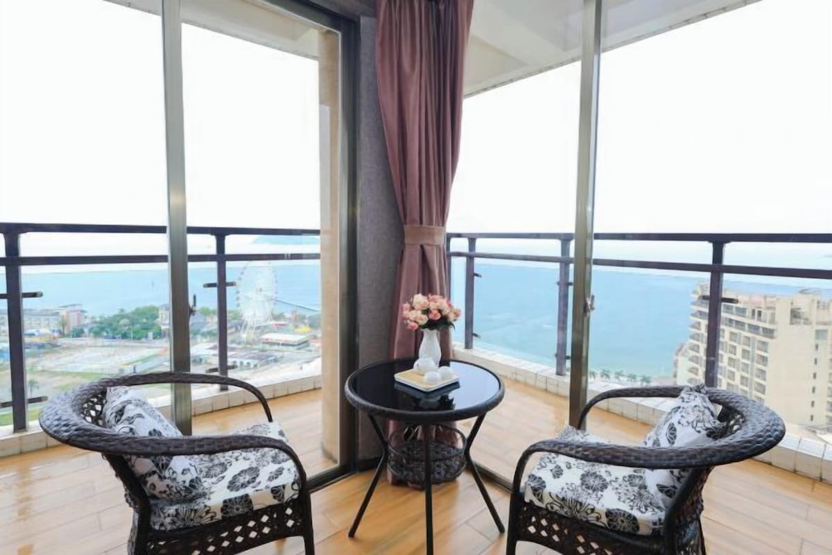 A balcony with two wicker chairs, a small round table with a vase of flowers, and a view of the sea and buildings.