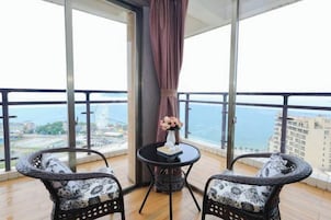 Un balcón con dos sillas de mimbre, una mesita redonda con un florero y vistas al mar y edificios.