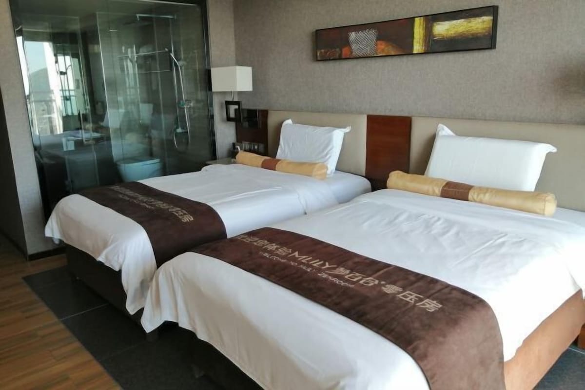 A hotel room with two beds, a glass shower enclosure, a wall-mounted lamp, and a painting.