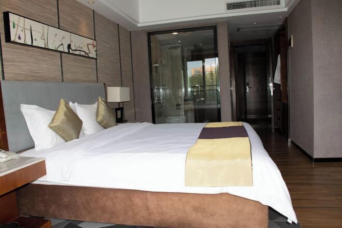 A hotel room with a large bed, a nightstand, a wall-mounted artwork, and a sliding glass door leading to a hallway.