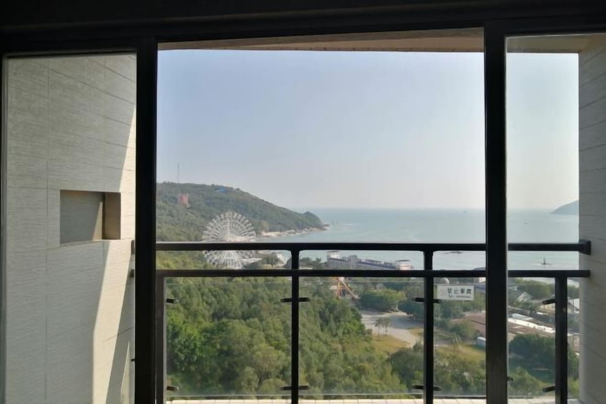 A view from a window with metal bars, overlooking a coastal area with a Ferris wheel and greenery.