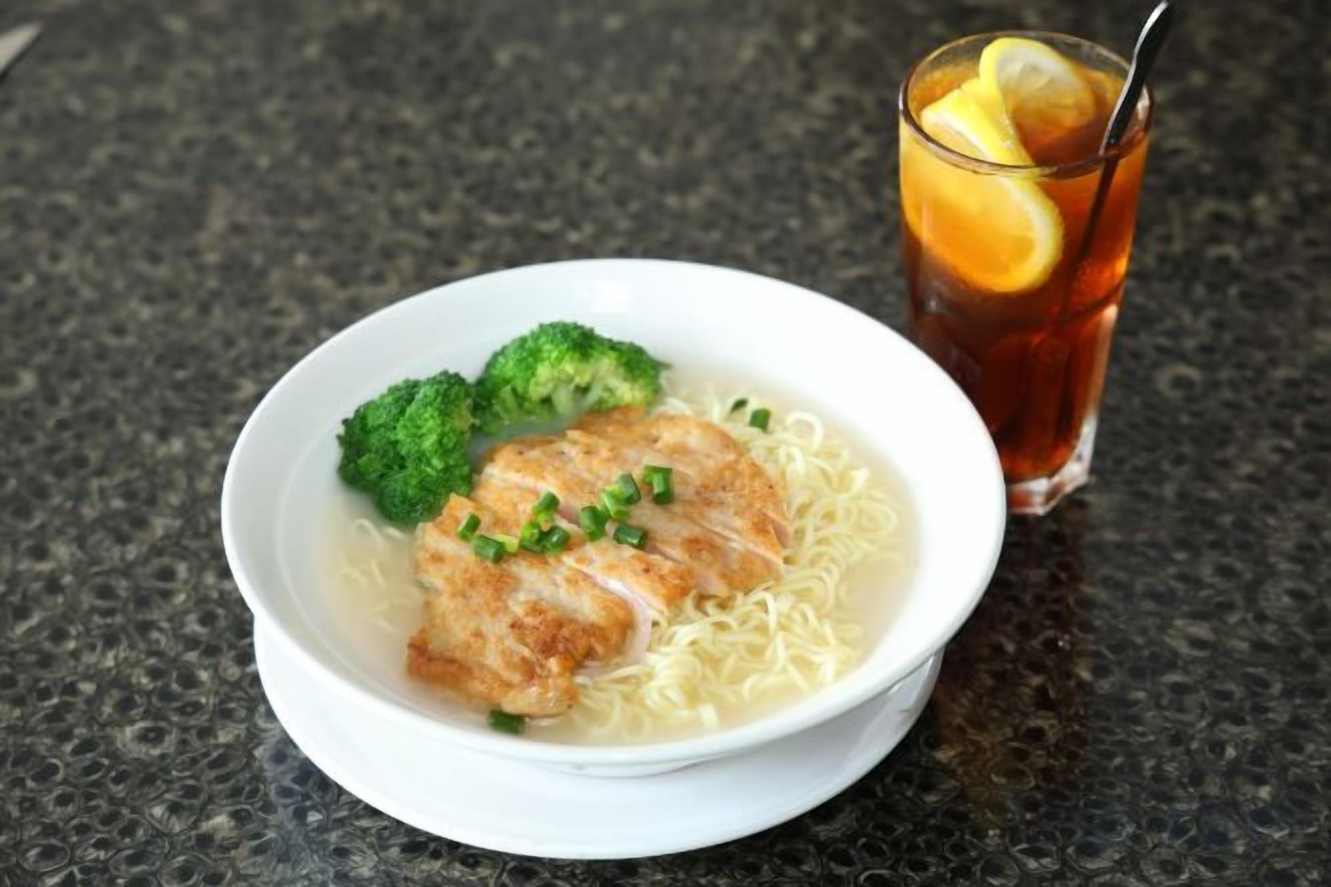 A bowl of ramen with a piece of chicken, broccoli, and a glass of iced tea with a lemon slice on a dark countertop.