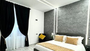 A modern bedroom with a large bed, a nightstand, a chair, and a window with black curtains. - Turati Inn (Rome)