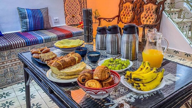 A dining table with various breakfast items, a sofa with colorful cushions, and a decorative wall with a geometric pattern. - Cozy Ensuite Room with AC

 (Tangier)
