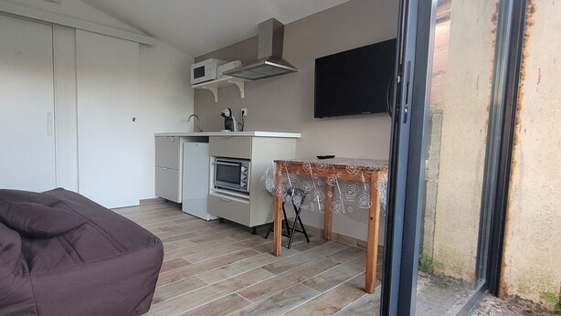 Una cocina compacta con microondas, horno y lavamanos, una pequeña mesa de comedor con un mantel decorativo y un televisor montado en la pared.