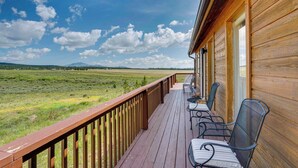 Una terraza de madera con mobiliario exterior, con vistas a un campo de hierba y montañas lejanas.