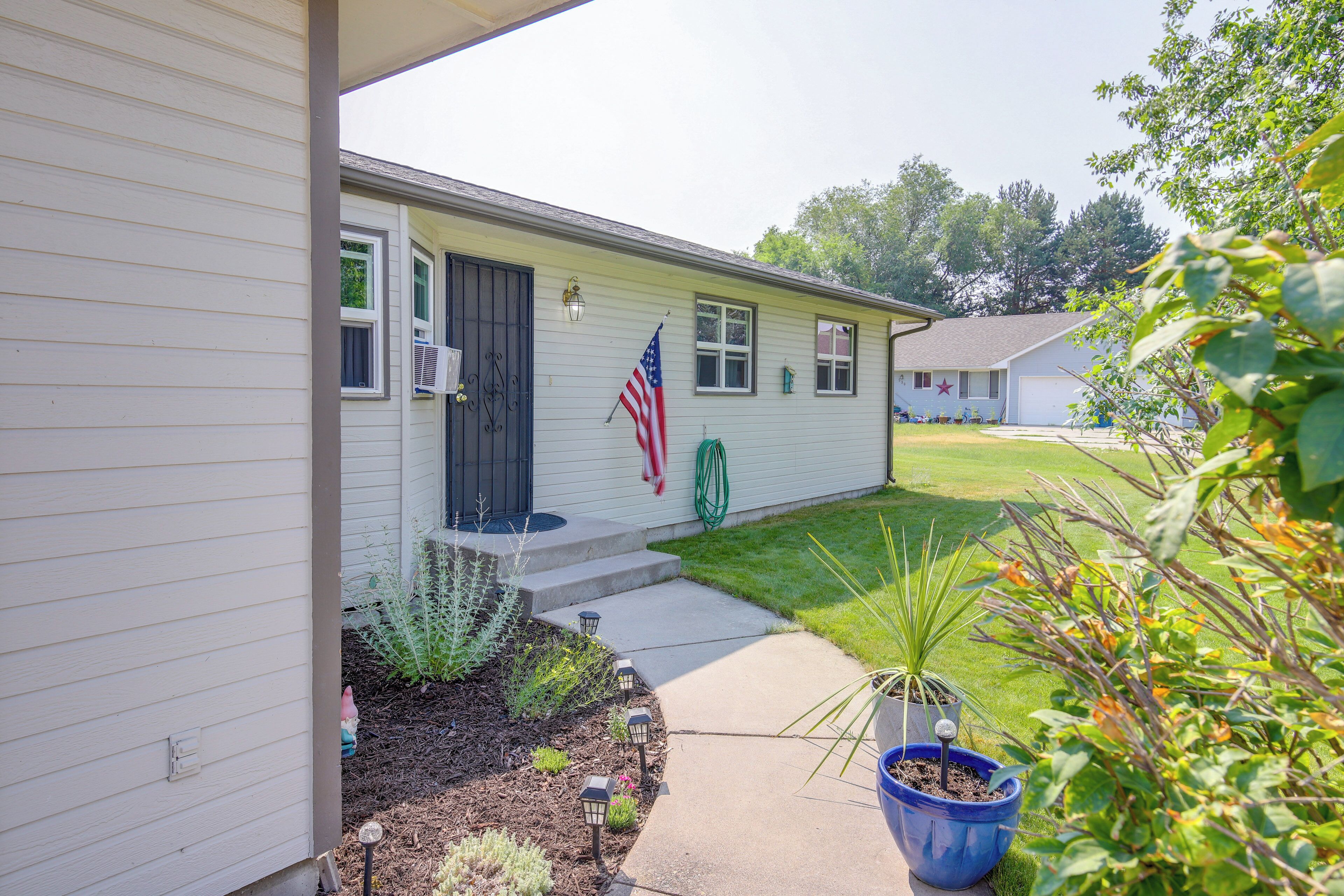 A single-story house with a front porch, a small garden with various plants, and an American flag.