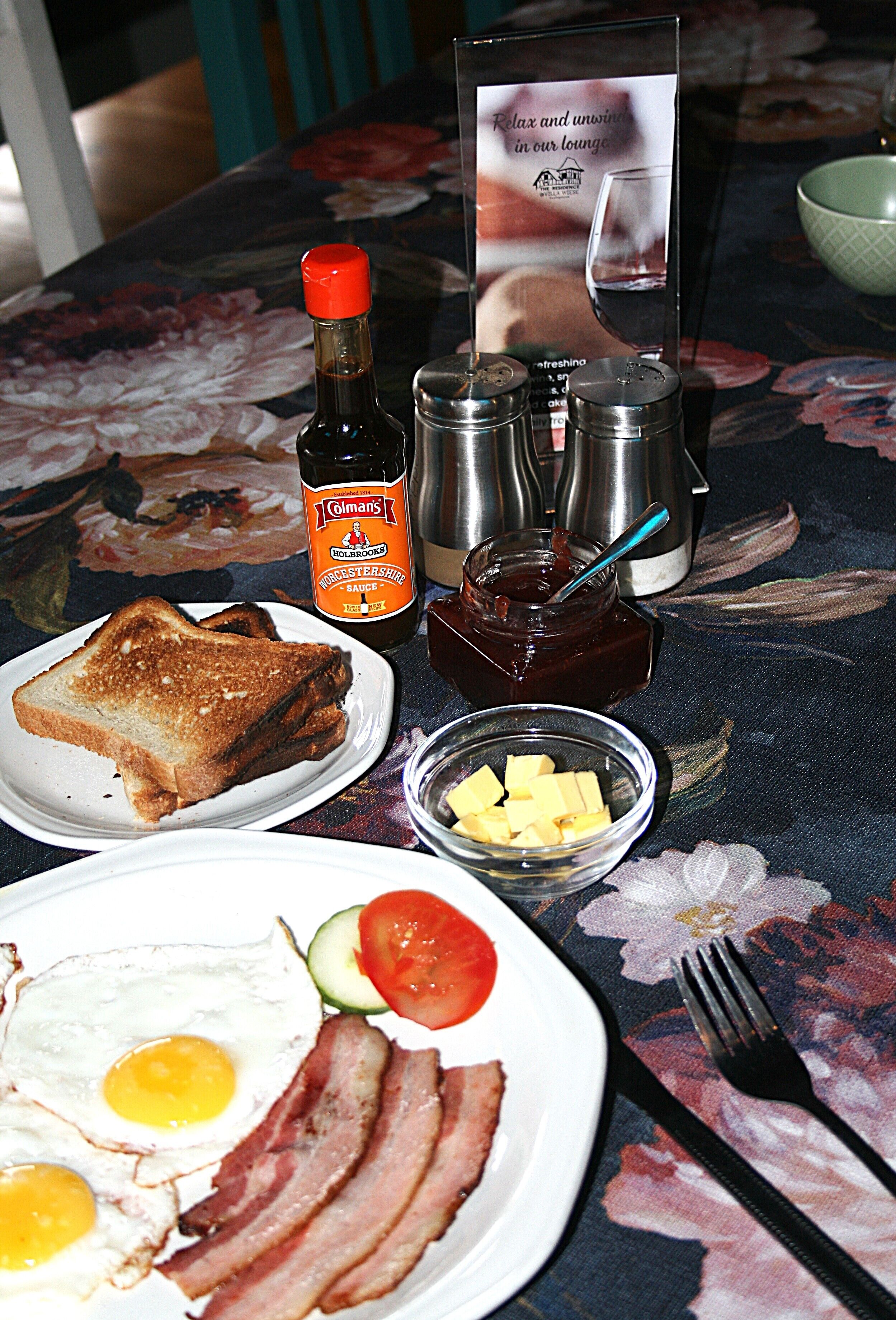 A table with a floral tablecloth, a bottle of Worcestershire sauce, condiments, a bowl of butter cubes, a plate with toast, eggs, bacon, and a tomato.