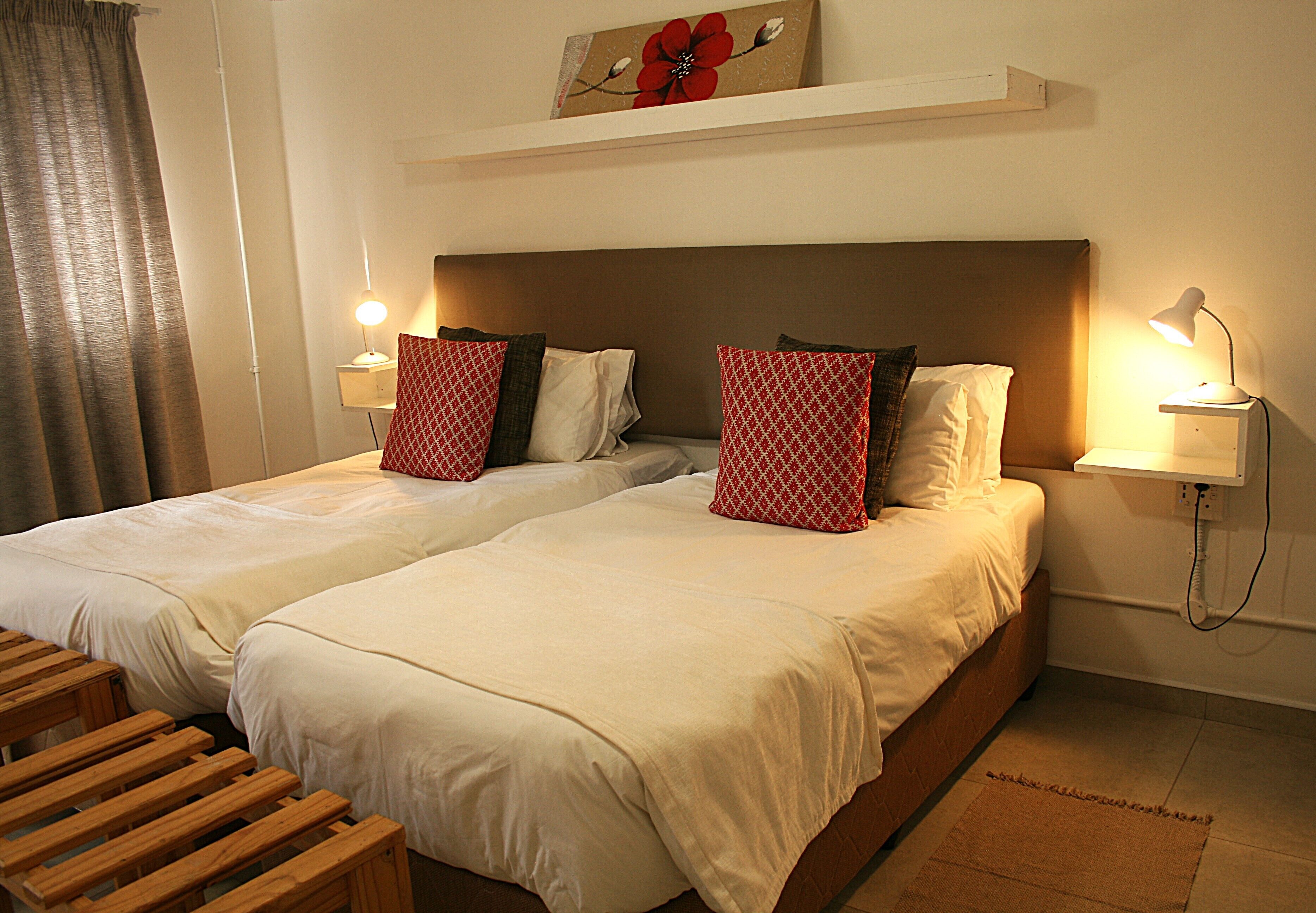 Ein Hotelzimmer mit zwei Betten, jeweils mit weißer Bettwäsche und roten Kissen mit Muster. An den Betten stehen Nachttische mit Lampen, eine hölzerne Bank und ein an der Wand befestigtes Regal.