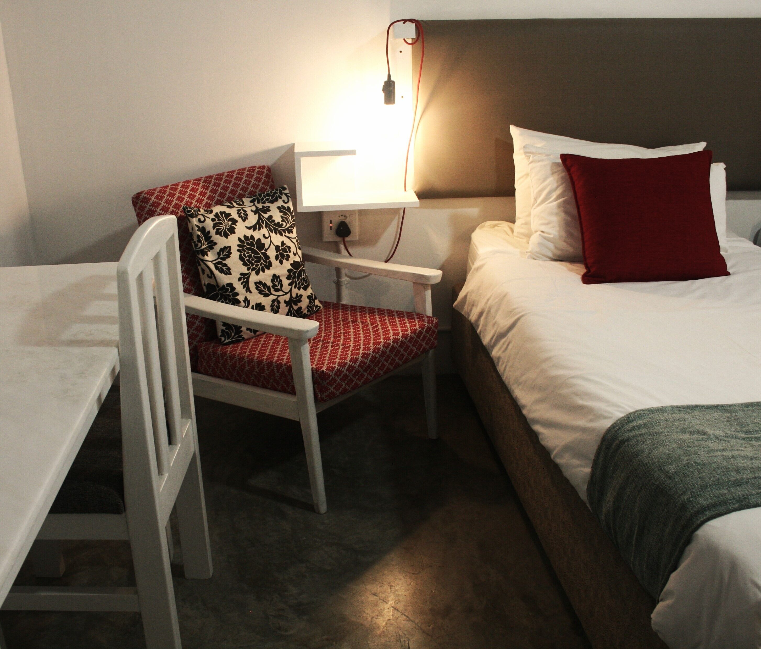 Ein Hotelzimmer mit Bett, Stuhl mit gemustertem Kissen, Nachttisch und Beistelltisch mit Lampe.